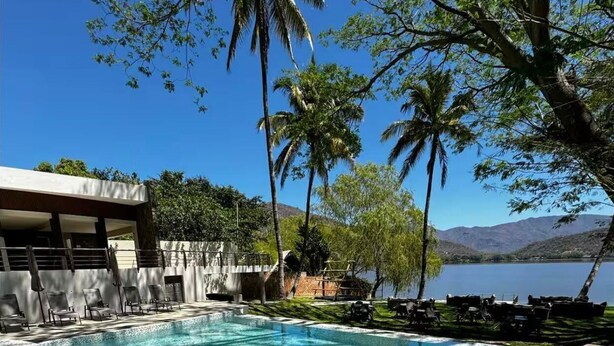 Una piscina con agua azul cristalina, rodeada de palmeras y un edificio con área de estar al aire libre.