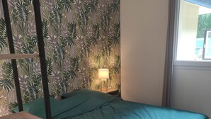 A room with a bed, a nightstand with a lamp, a window, and a wall with a leaf pattern. - L'Appartement Myrade, pretty T2 on the edge of the forest (Biscarrosse)