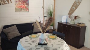 A cozy living room with a dark-colored sofa, a round dining table with a patterned tablecloth, and a cabinet with decorative items. - L'Appartement Myrade, pretty T2 on the edge of the forest (Biscarrosse)