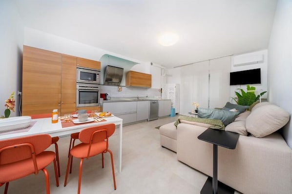 Een moderne keuken met witte kasten, een eethoek met oranje stoelen, en een woonkamer met een beige bank en een flatscreen televisie.