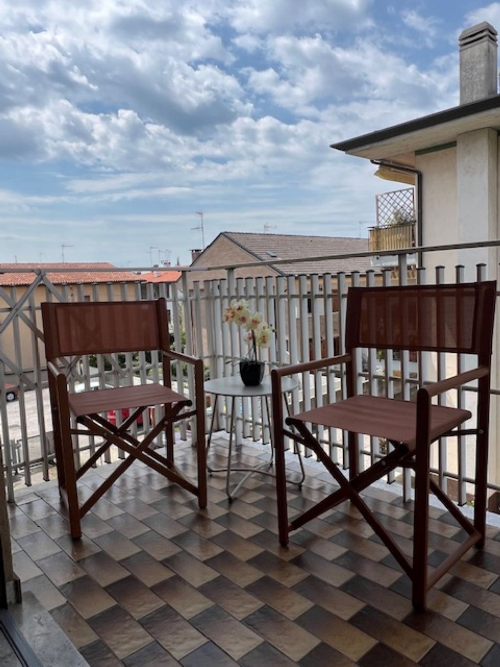 Eine Terrasse mit zwei Holzstühlen, einem Tisch, einem Blumentopf und Blick auf den Himmel und Gebäude.