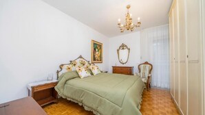 Quarto com piso de madeira, colcha verde, lustre, mesa de cabeceira de madeira, uma cadeira e um guarda-roupa.