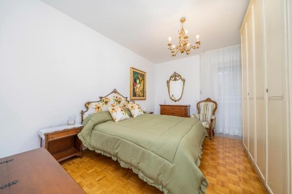 Une chambre avec un plancher en bois, une couvre-lit verte, un lustre, une table de chevet en bois, une chaise et une armoire.