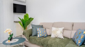 Sala de estar com um sofá bege, manta verde e almofadas estampadas. Uma pequena mesa redonda com toalha de renda branca e uma orquídea é visível. Uma planta verde grande e uma orquídea em vaso estão sobre o sofá.