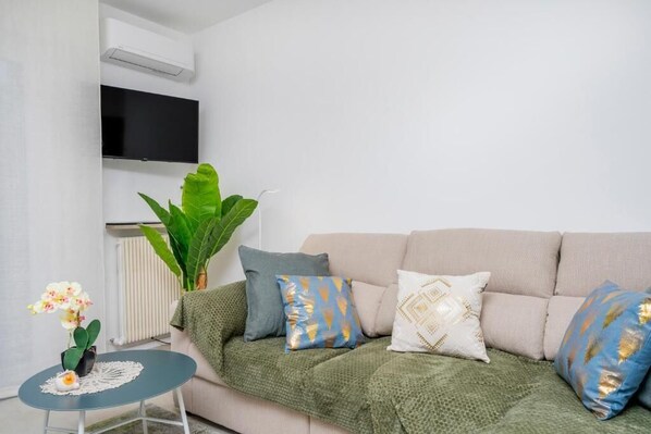 A living room with a beige sofa, green throw blanket, and patterned pillows. A small round table with a white lace tablecloth and an orchid plant is visible. A large green plant and a potted orchid are placed on the sofa. - MIRELLA's cute place - Udine Italy (Udine)