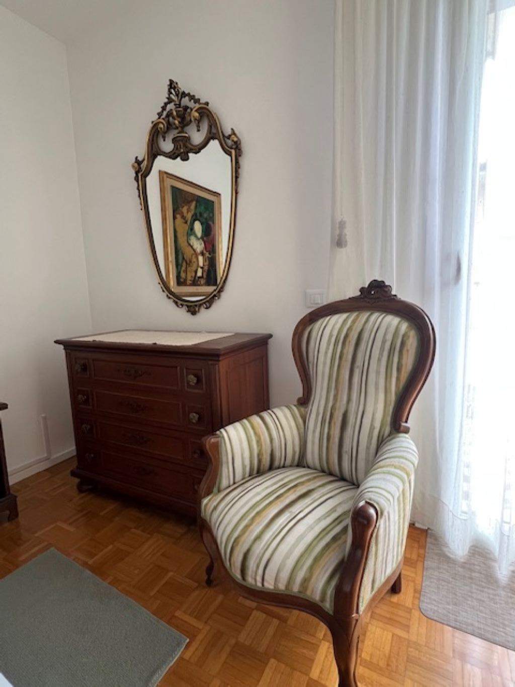 Una habitación con tocador de madera, un sillón a rayas, un cuadro en la pared y una ventana con cortinas.