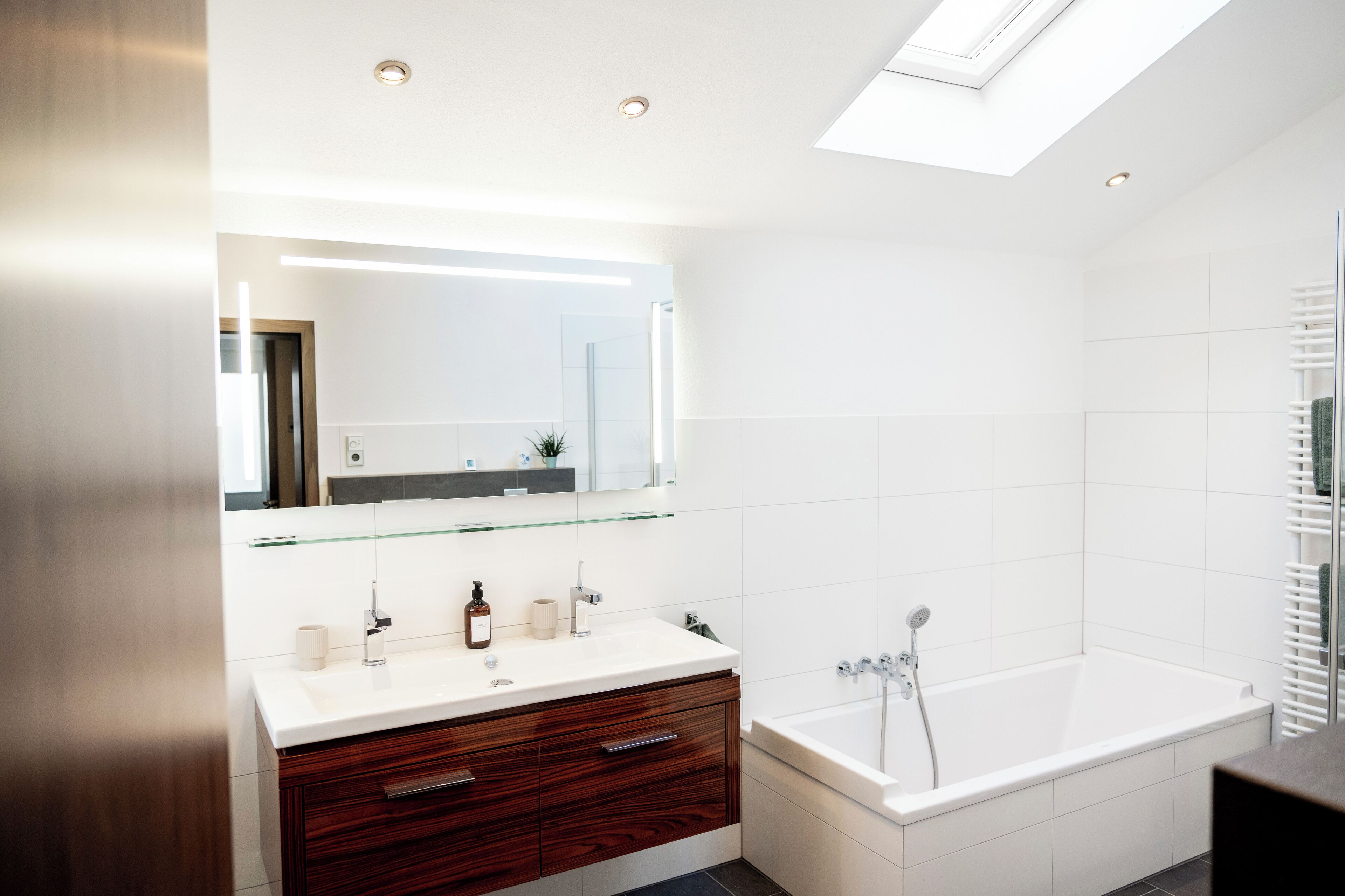 Une salle de bain moderne avec une lucarne, des carreaux blancs, un meuble-lavabo en bois, une baignoire blanche et une étagère en verre au-dessus du lavabo.