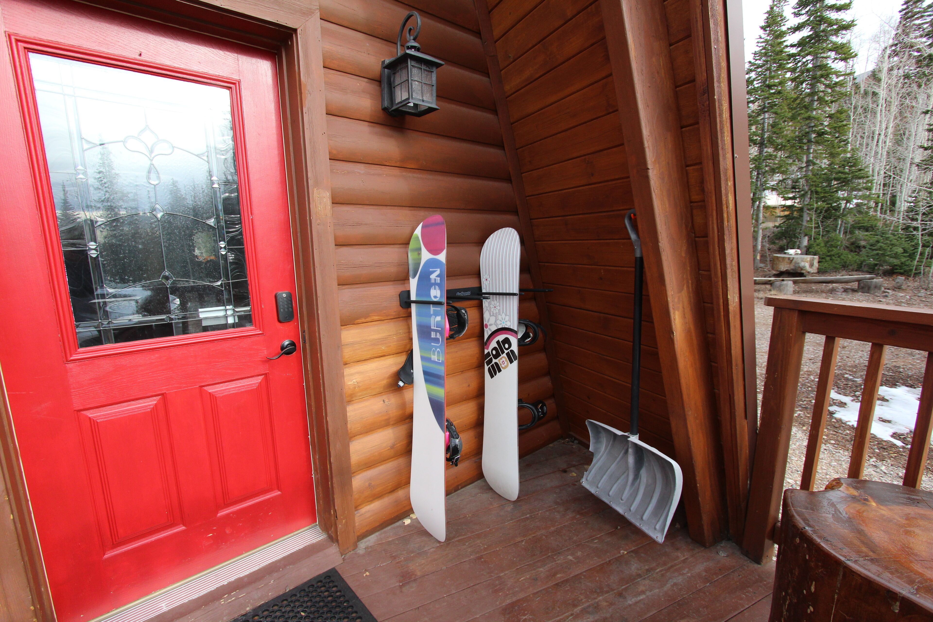 A wooden cabin with a red door, two snowboards leaning against the wall, and a shovel on the floor.