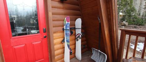 A wooden cabin with a red door, two snowboards leaning against the wall, and a shovel on the floor.