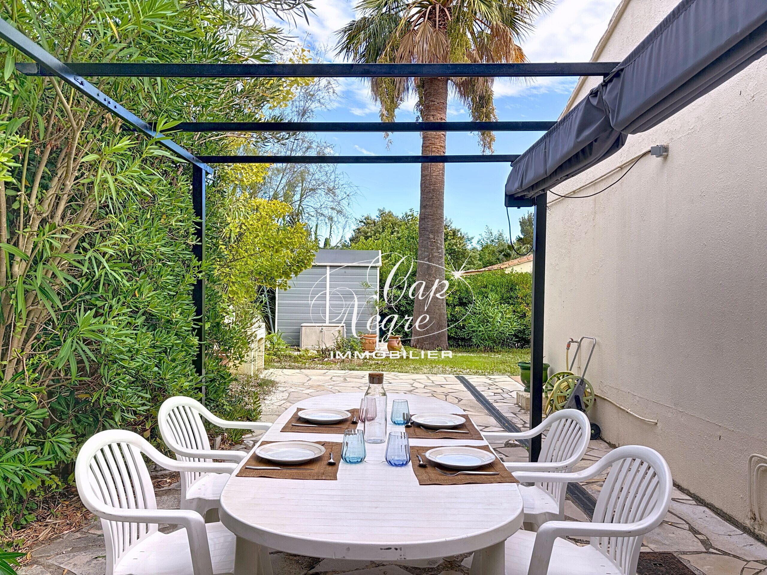 Een overdekt buitenDininggedeelte met een tafel voor vier personen, omringd door witte stoelen, onder een metalen pergola.