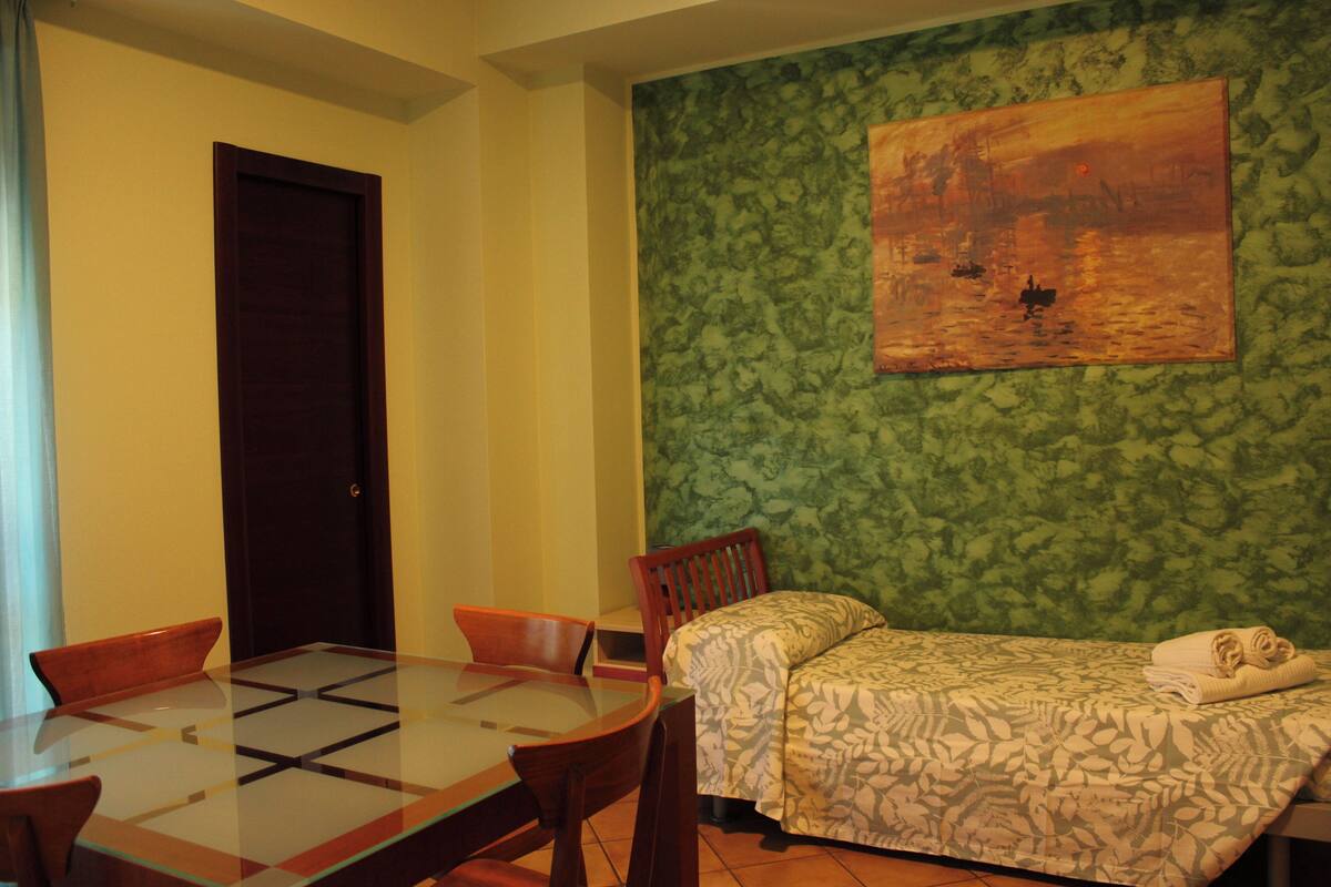A room with a glass-top dining table, wooden chairs, a bed with patterned bedding, and a green textured wall.