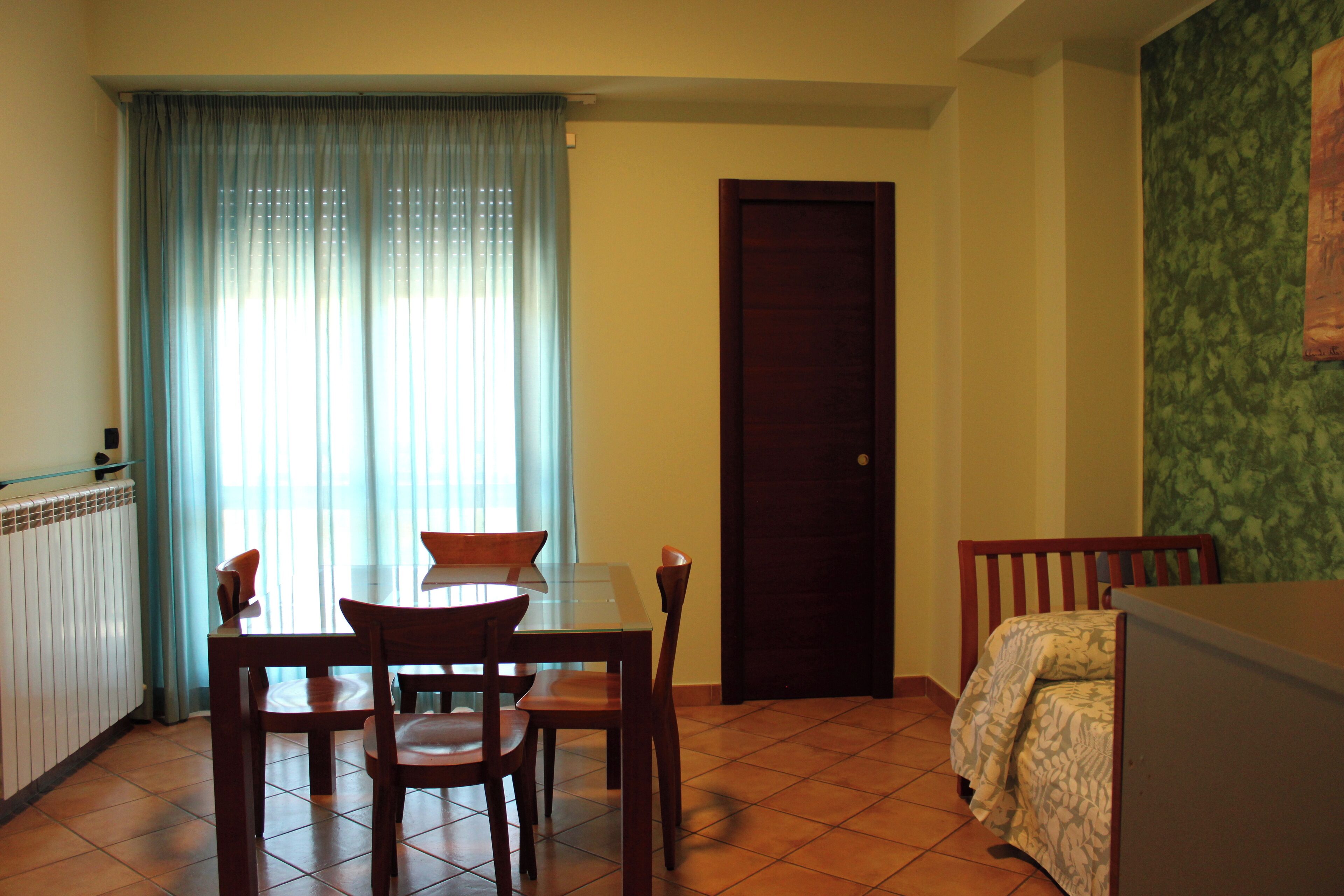 A dining area with a table and chairs, a window with curtains, a door, and a bed with a patterned cover.
