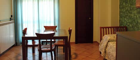 A dining area with a table and chairs, a window with curtains, a door, and a bed with a patterned cover.
