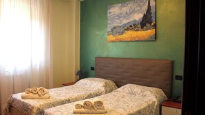 Una camera d'albergo con due letti, ognuno con una coperta a fantasia e asciugamani arrotolati. Sopra i letti, un quadro appeso al muro.