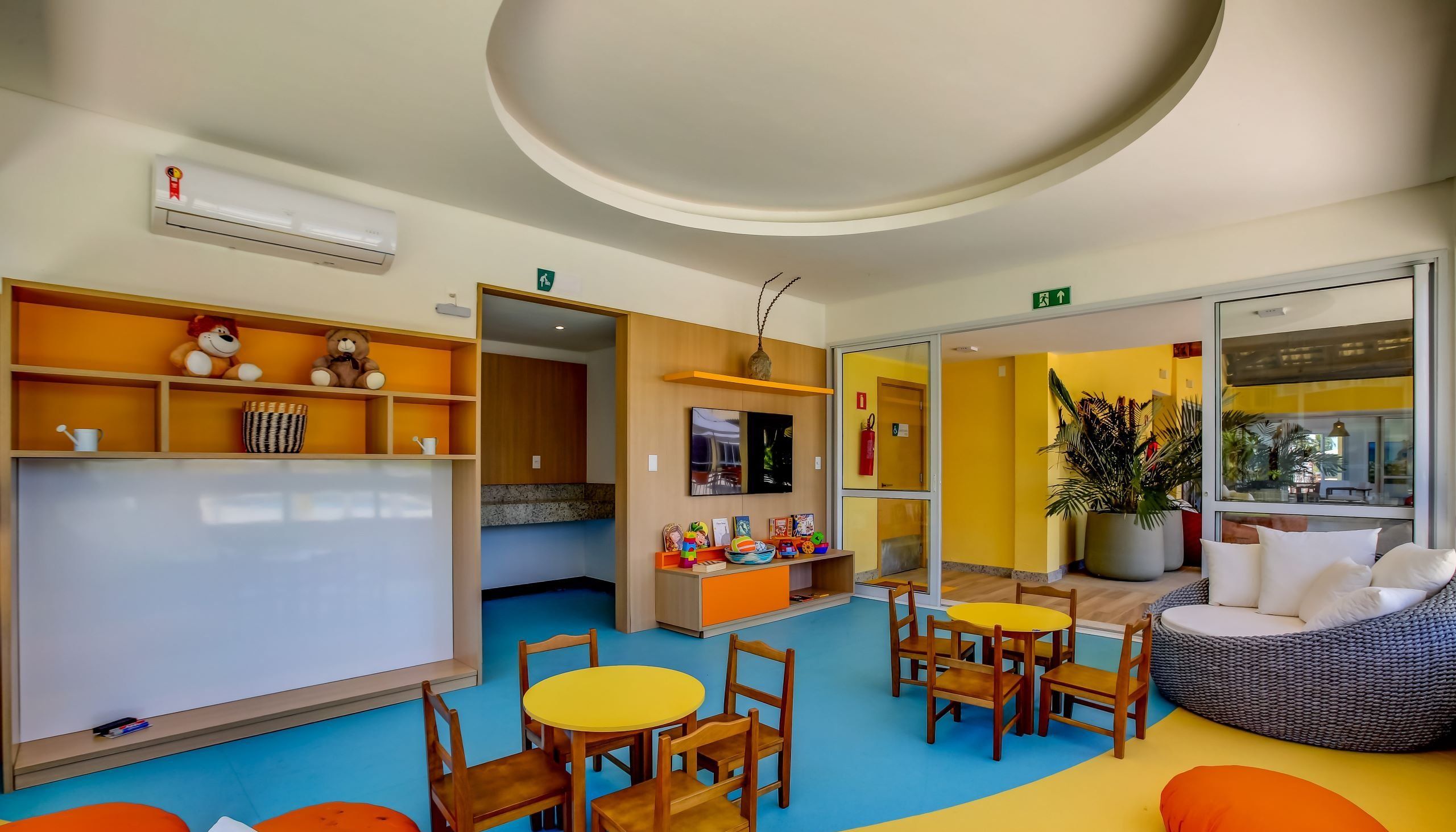 Uma área de recreação infantil com mobiliário colorido, um quadro branco e uma planta grande.