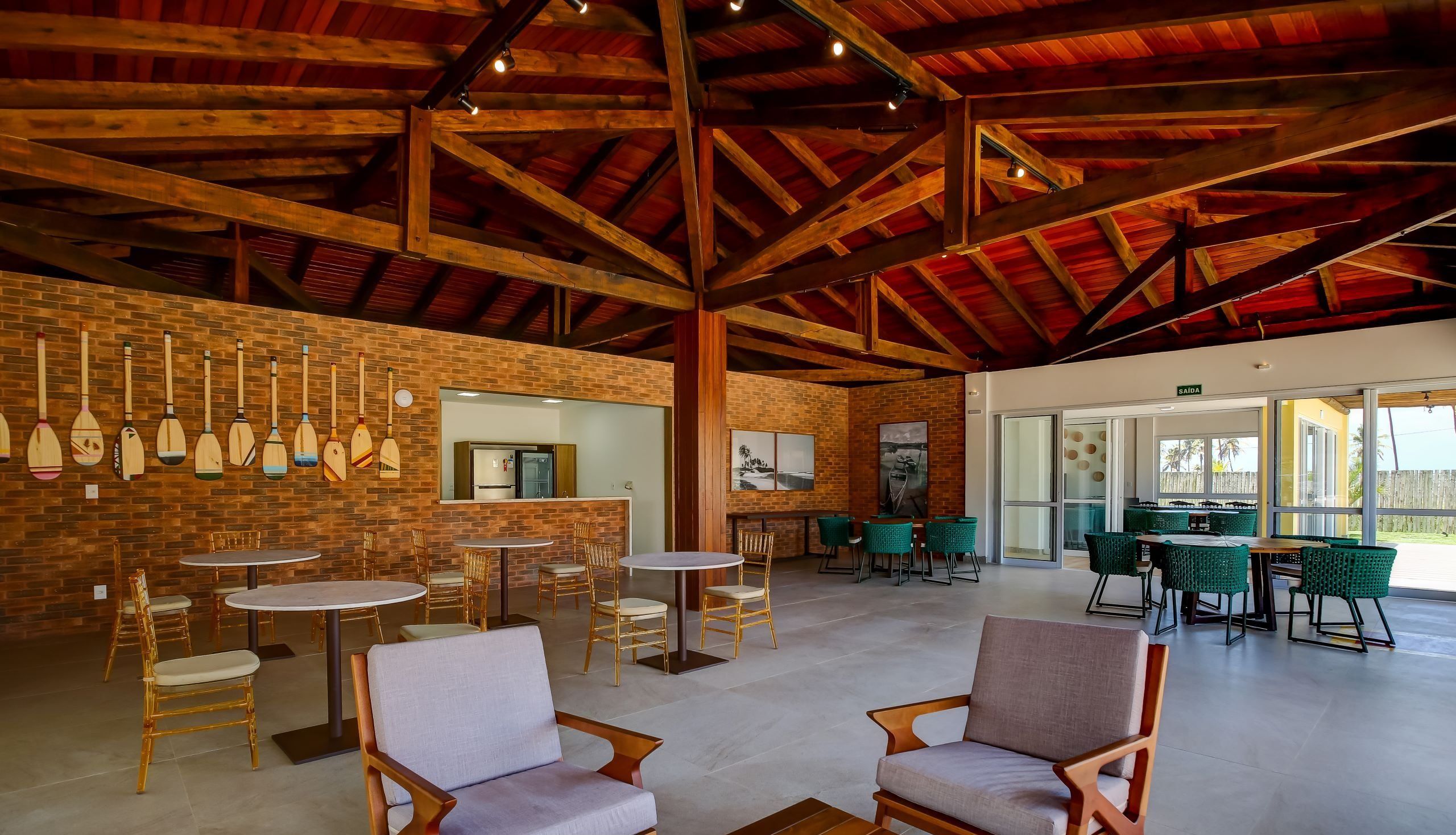 A spacious indoor area with high ceilings and exposed wooden beams, featuring multiple seating arrangements and a brick wall with paddleboards displayed.
