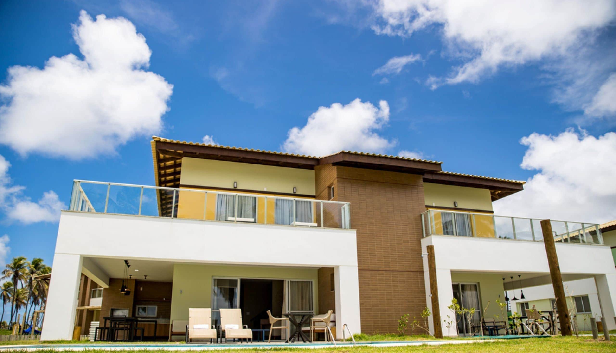 Un edificio de dos pisos con piscina, mobiliario exterior y cielo azul despejado.