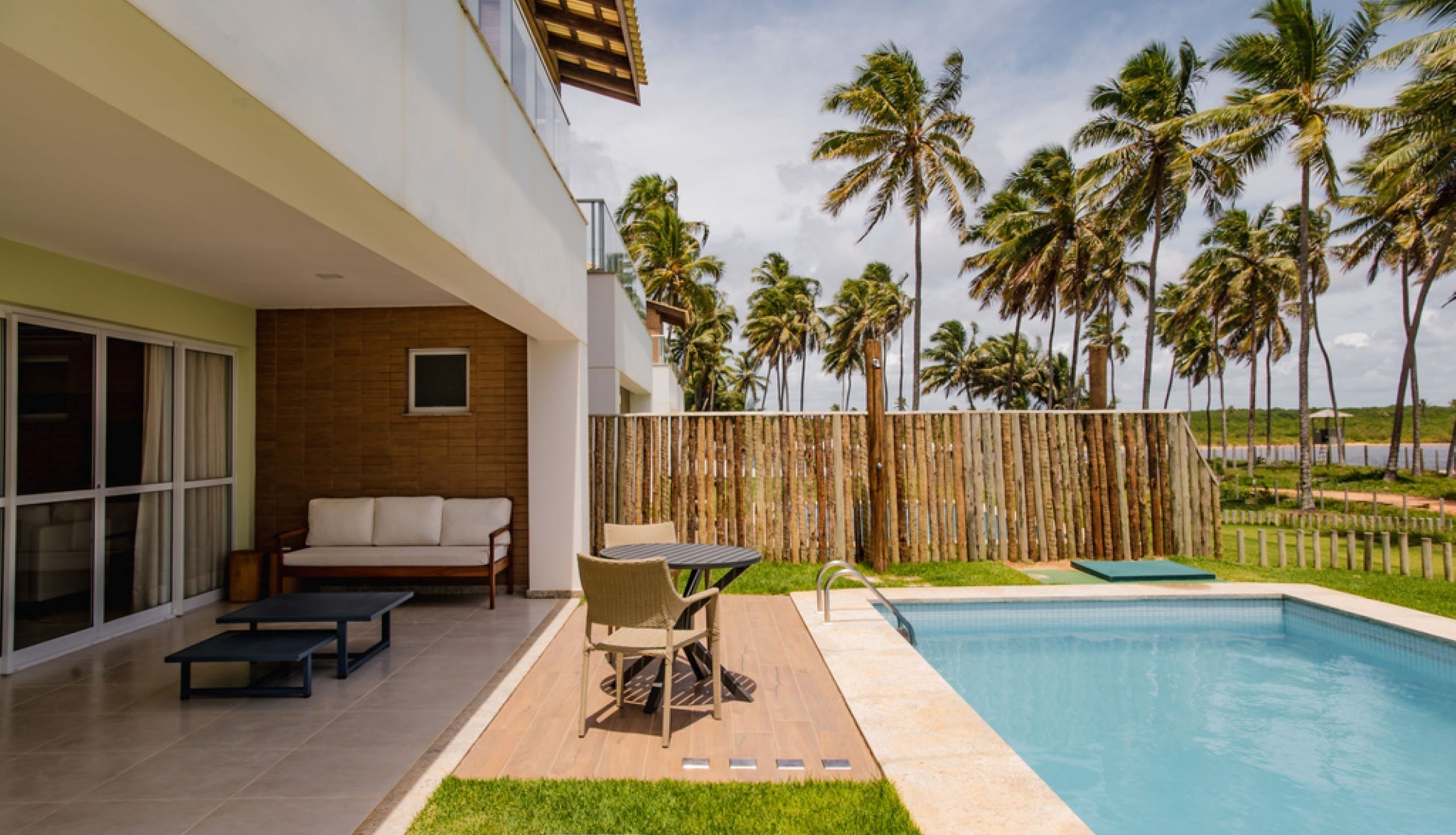 A modern outdoor living area with a pool, a wooden fence, and palm trees in the background.