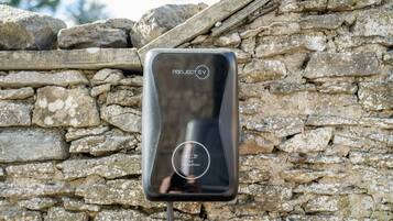 Uma estação de carregamento para veículo elétrico preto, com a inscrição "PROJECT EV", em frente a uma parede de pedra.