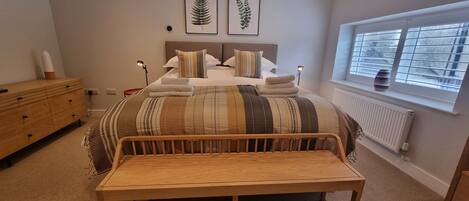 A bedroom with a bed, bedside lamps, a wooden bench, a dresser, and a window with blinds.