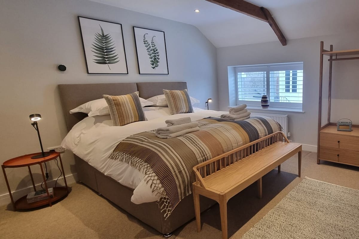 A bedroom with a bed, a bench, a bedside table, a radiator, a window with blinds, and a coat rack.