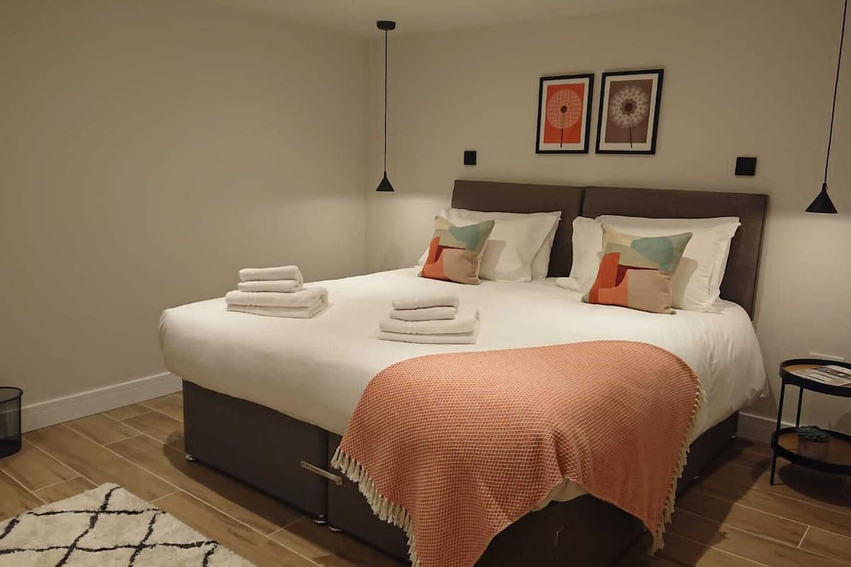 A neatly made bed with white linens and a peach-colored throw blanket, a small bedside table with a magazine, and two wall-mounted lamps.