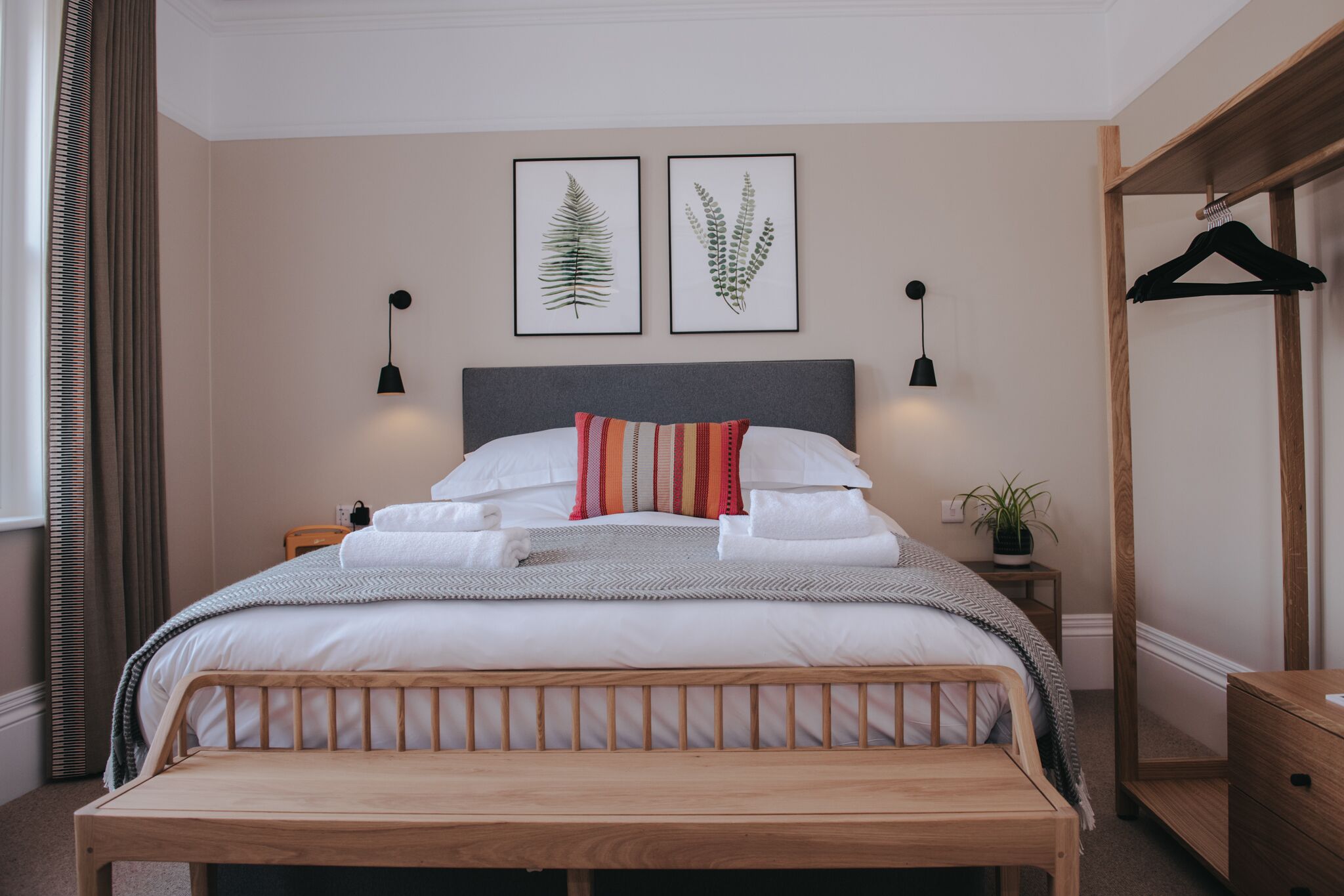 A neatly made bed with white linens and a striped pillow, a bedside table with a plant, and two framed botanical prints on the wall.