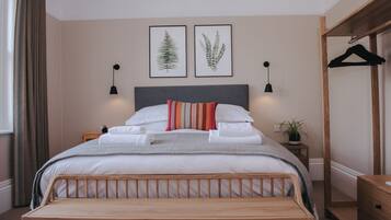 A neatly made bed with white linens and a striped pillow, a bedside table with a plant, and two framed botanical prints on the wall.