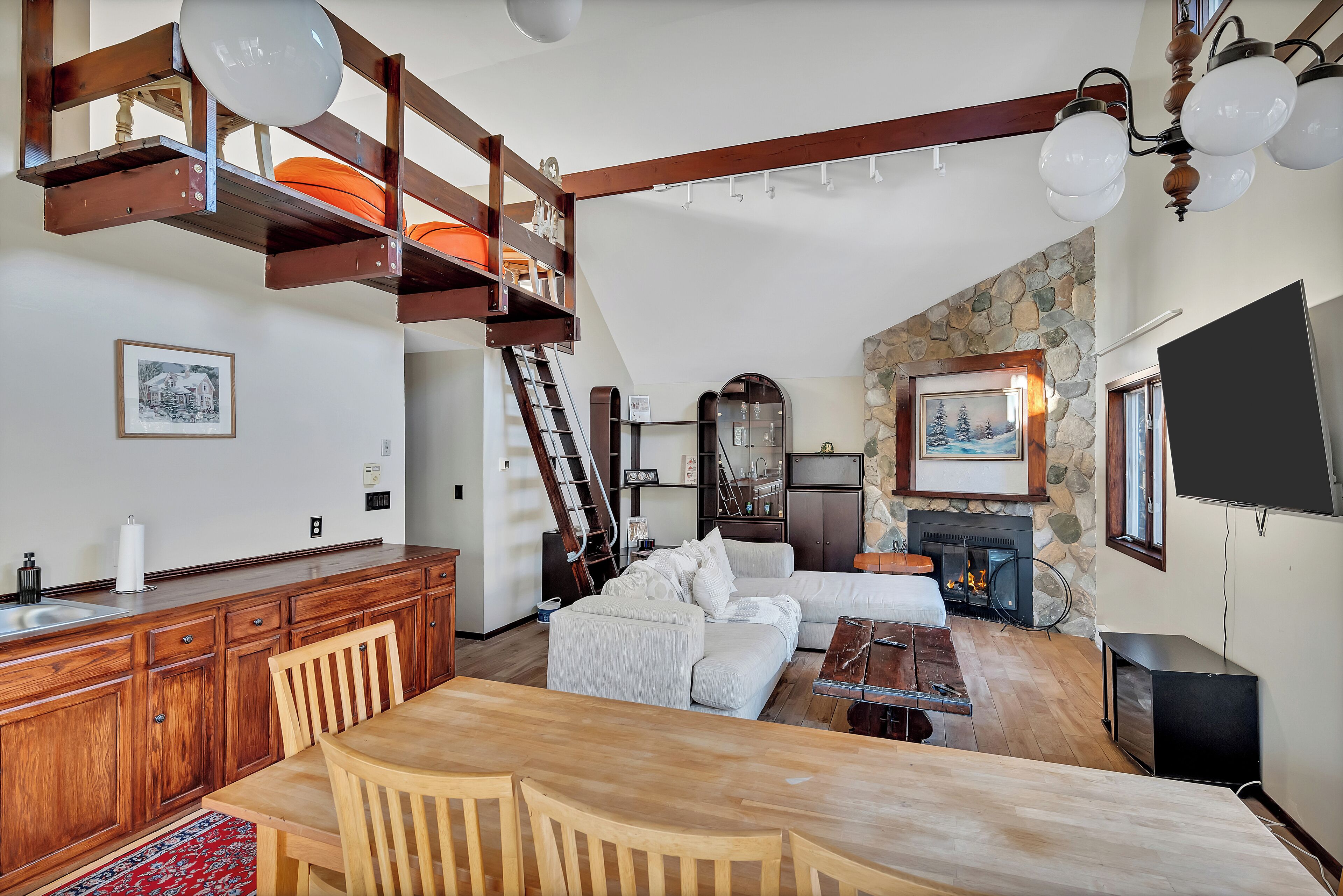 Ein geräumiger Wohnbereich mit einem Holzesstisch, einer Couch und einem Kamin. Über dem Wohnbereich befindet sich eine Galerie mit einer Treppe und einem Balkon.