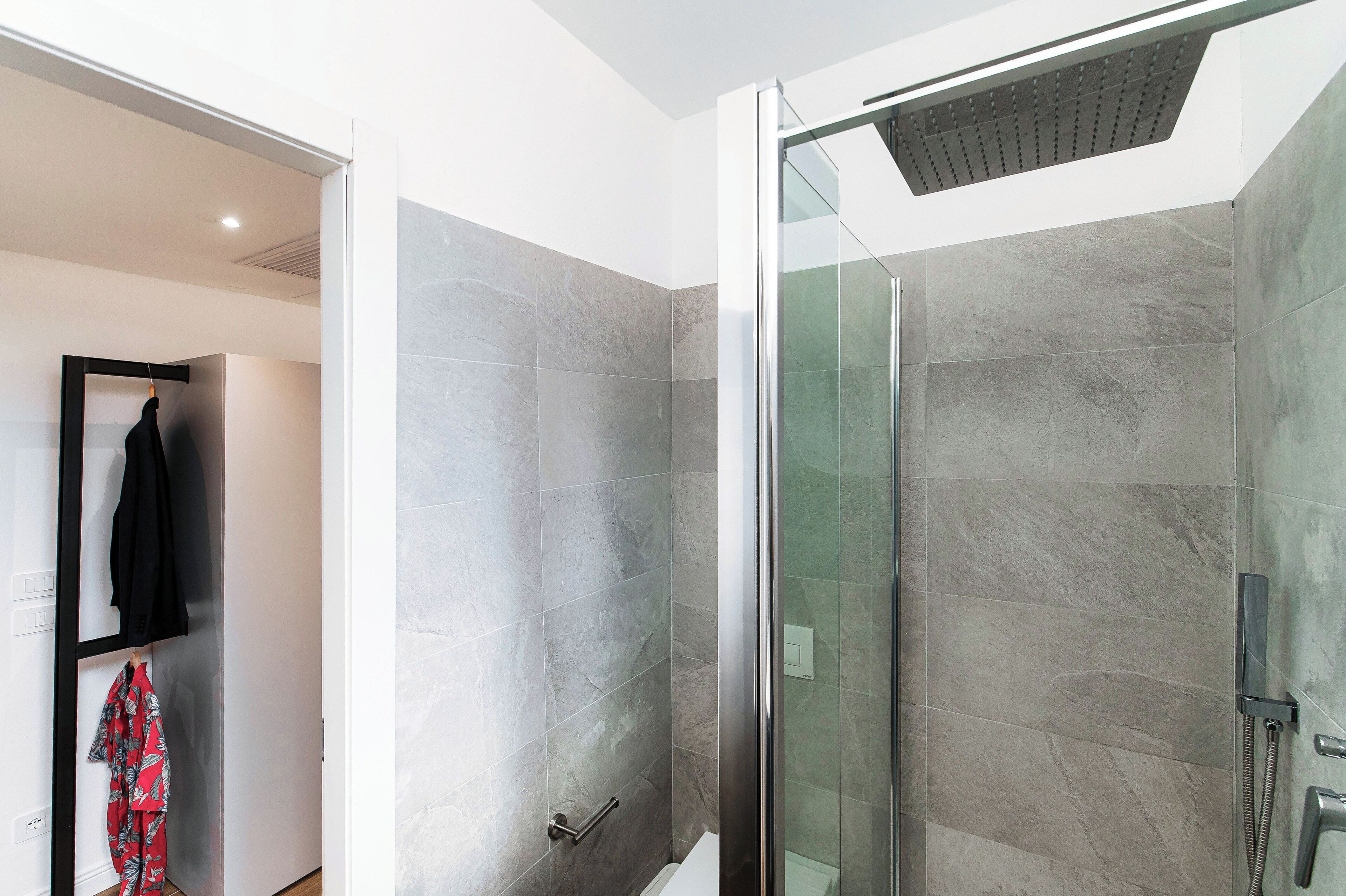 Ein modernes Badezimmer mit einer in Glas eingefassten Dusche, grauen Fliesen und einem Handtuchhalter mit einem schwarzen und einem roten, gemusterten Handtuch.