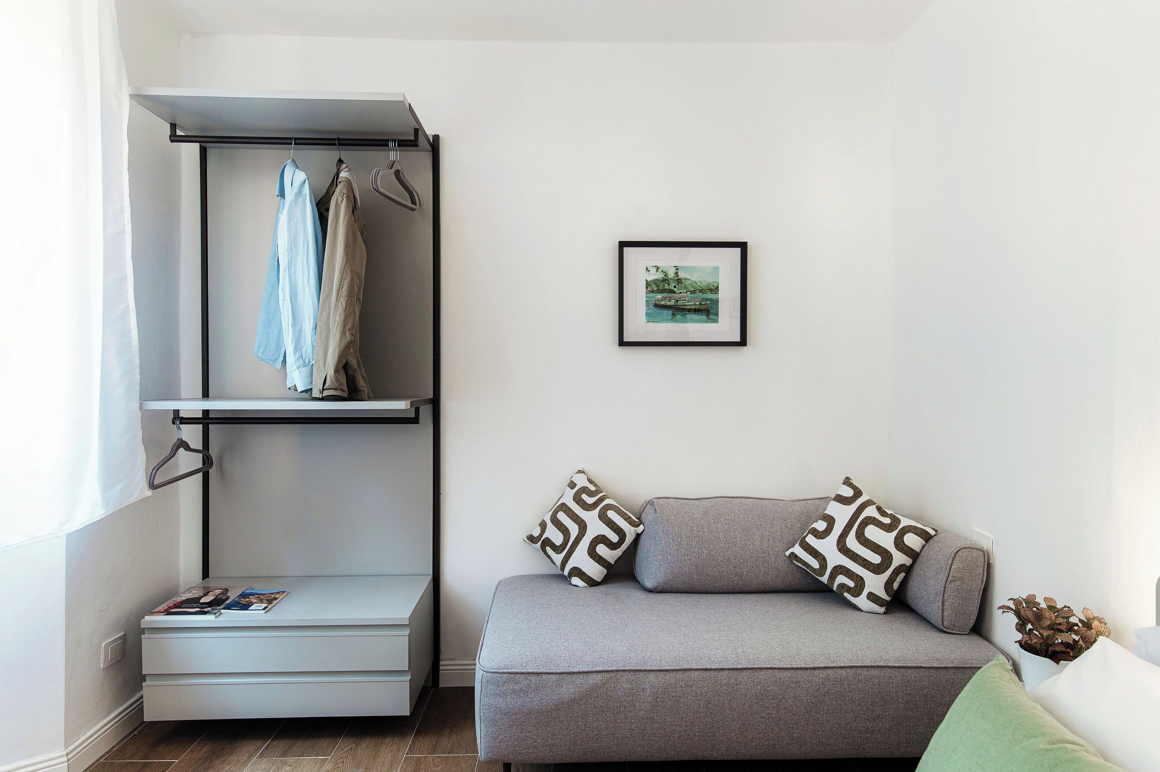 Ein kompakter Wohnraum mit grauem Sofa, gemusterten Kissen, einem kleinen Tisch mit Zeitschriften, einer Wandablage mit Kleidung und einem gerahmten Bild.