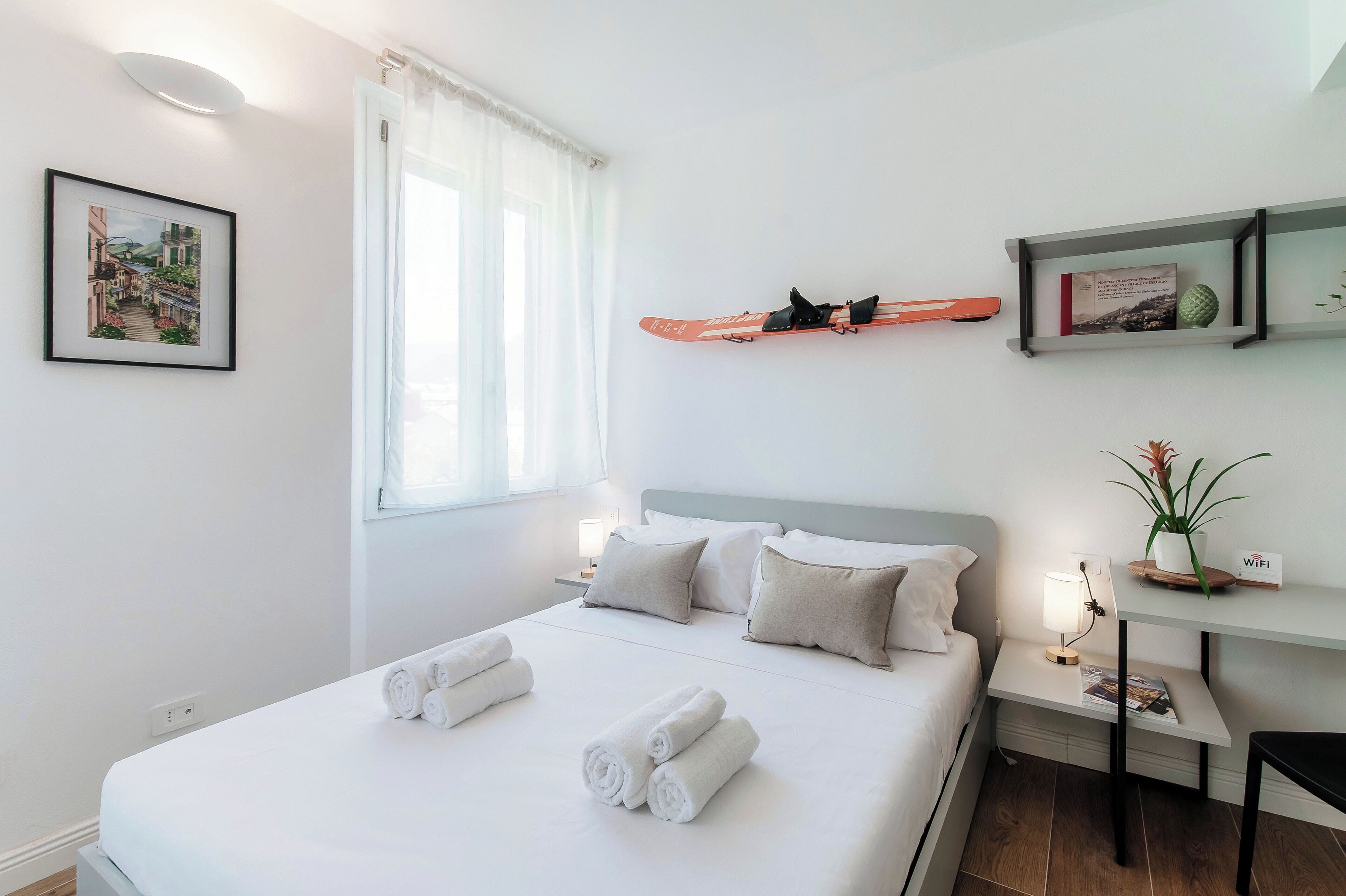 Una cama bien hecha con toallas enrolladas, una mesita de noche con una lámpara, un cuadro en la pared y un estante con un libro y un objeto decorativo.