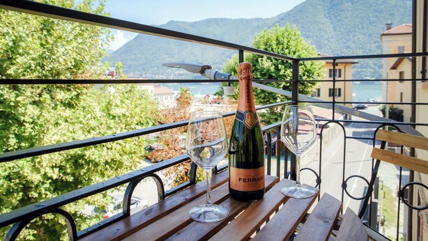 A balcony with a bottle of Ferrari wine and two glasses, overlooking a scenic lake and mountains. - Casa Batèl Suites & Apartments (Como)