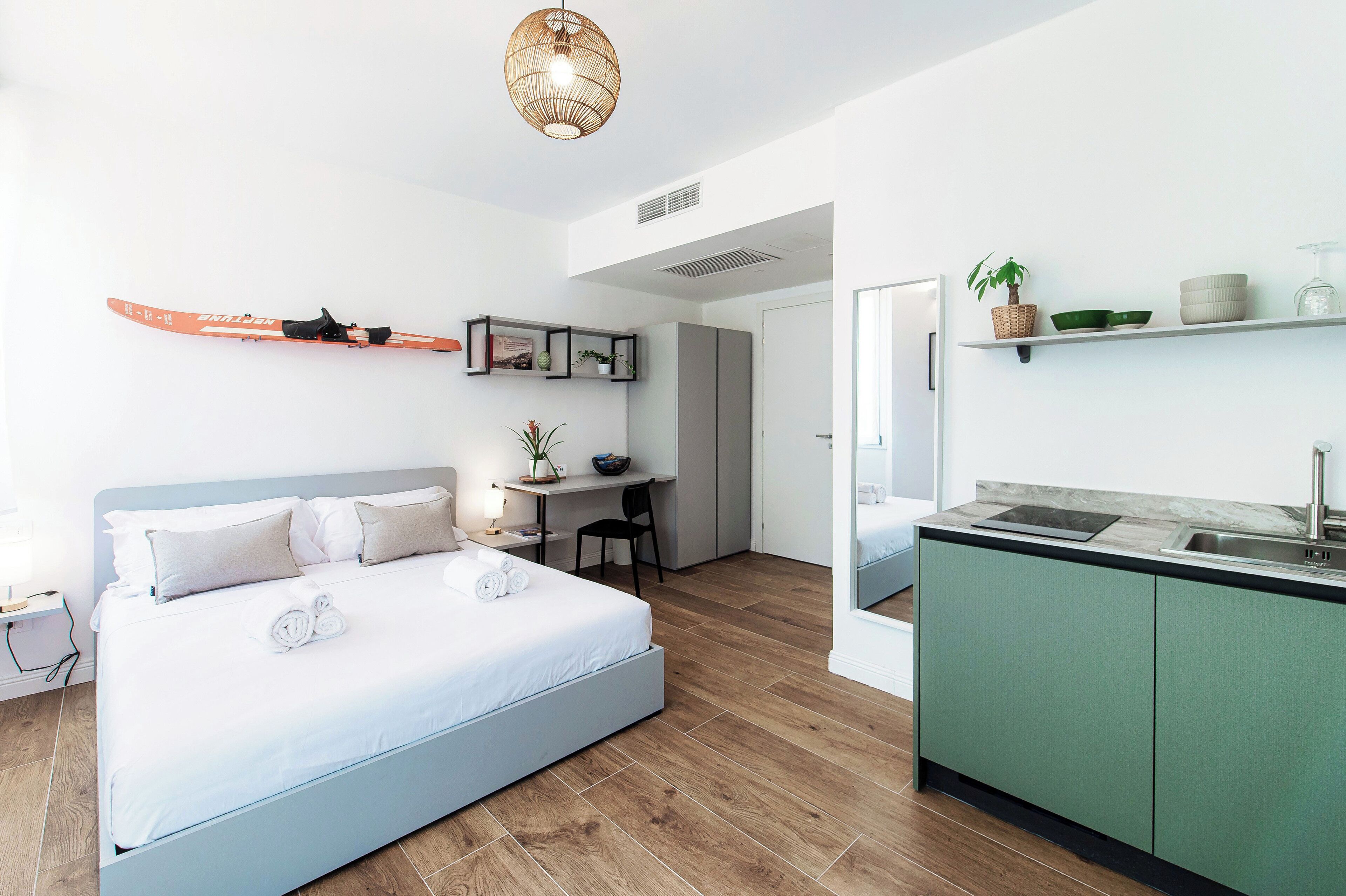 Ein modernes Schlafzimmer mit Bett, Nachttisch und Schreibtisch mit Stuhl. Es gibt eine Küchenzeile mit Spüle und Schränken.