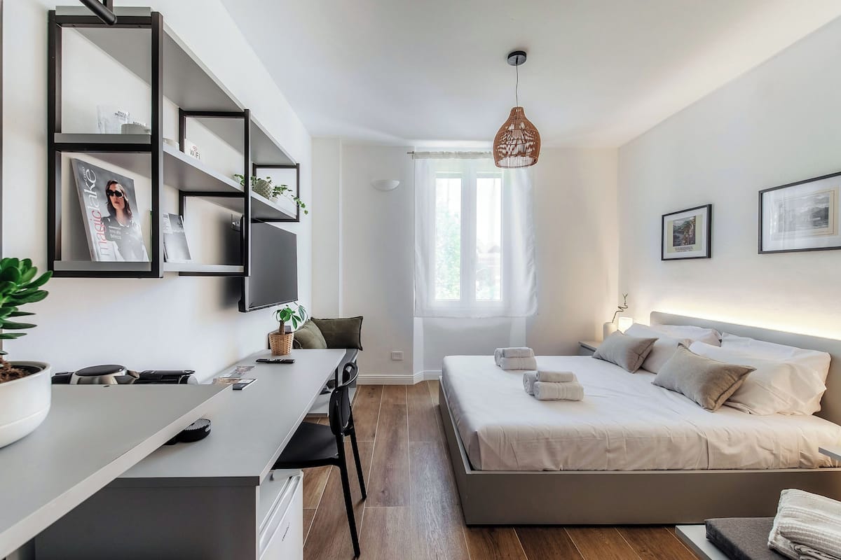 A modern bedroom with a bed, bedside table, and a desk with a chair. There is a window with curtains, a hanging light fixture, and a wall-mounted TV.