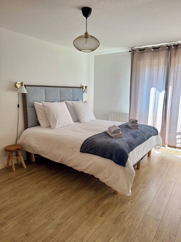 A bedroom with a bed, bedside lamps, a wooden stool, a radiator, and a window with curtains.