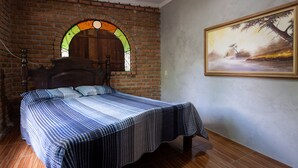 Un dormitorio con pared de ladrillo, una cama de madera, un cuadro y una ventana con diseño de vitral.