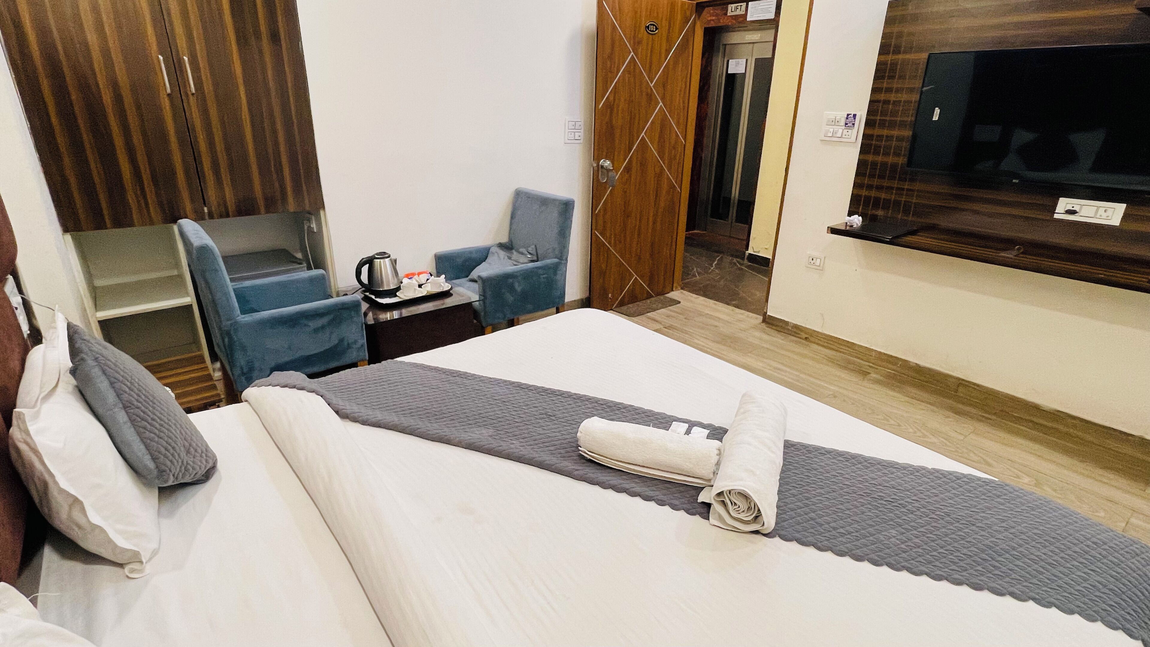 A hotel room with a bed, a desk with a kettle, a chair, a wardrobe, and a TV.