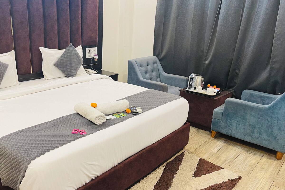 Una habitación de hotel con una cama grande, dos sillones, una mesita de noche con teléfono y cafetera, y una ventana con cortinas.