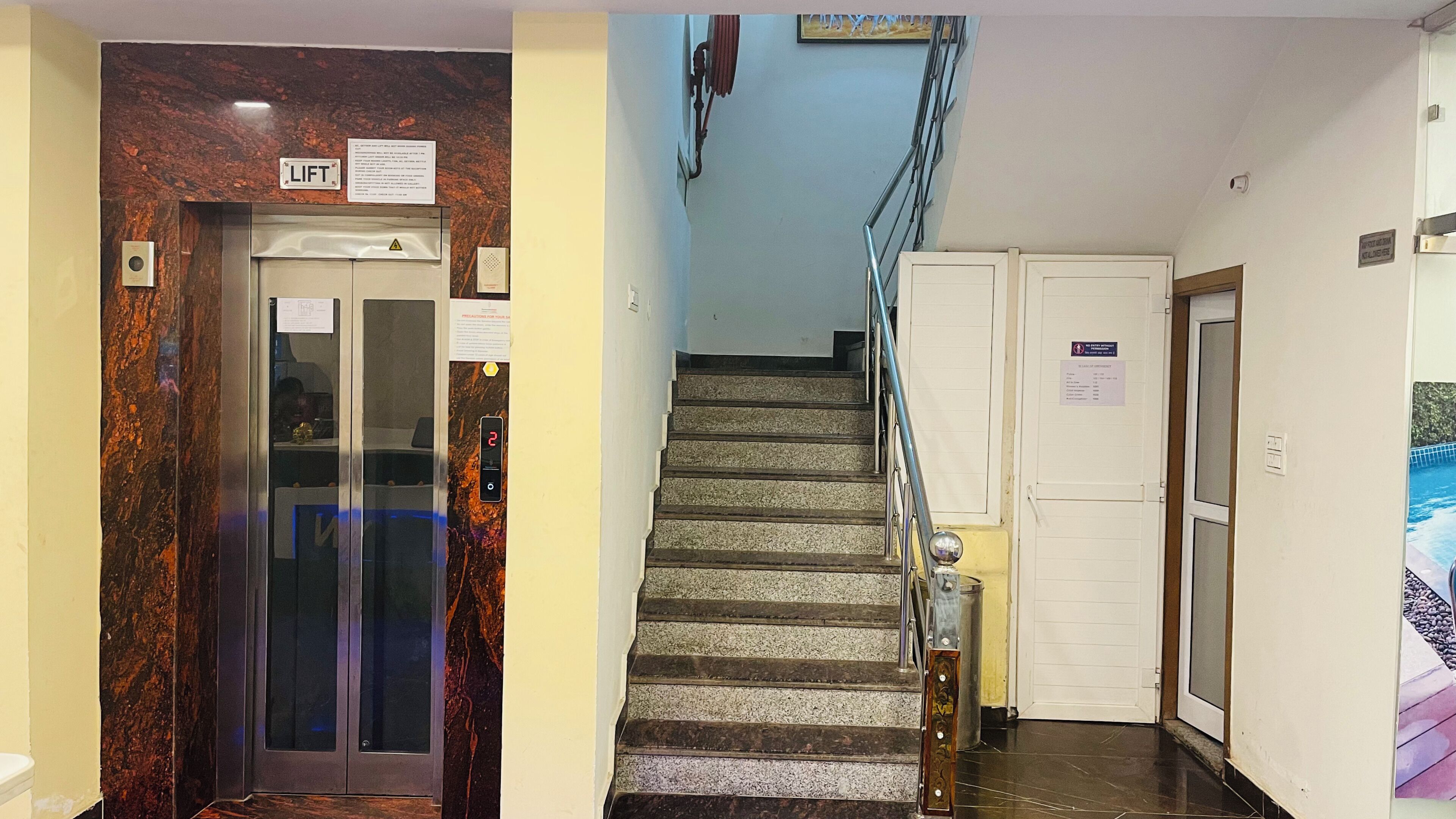 A hotel lobby with a marble elevator, a staircase with a handrail, and a door with a sign.
