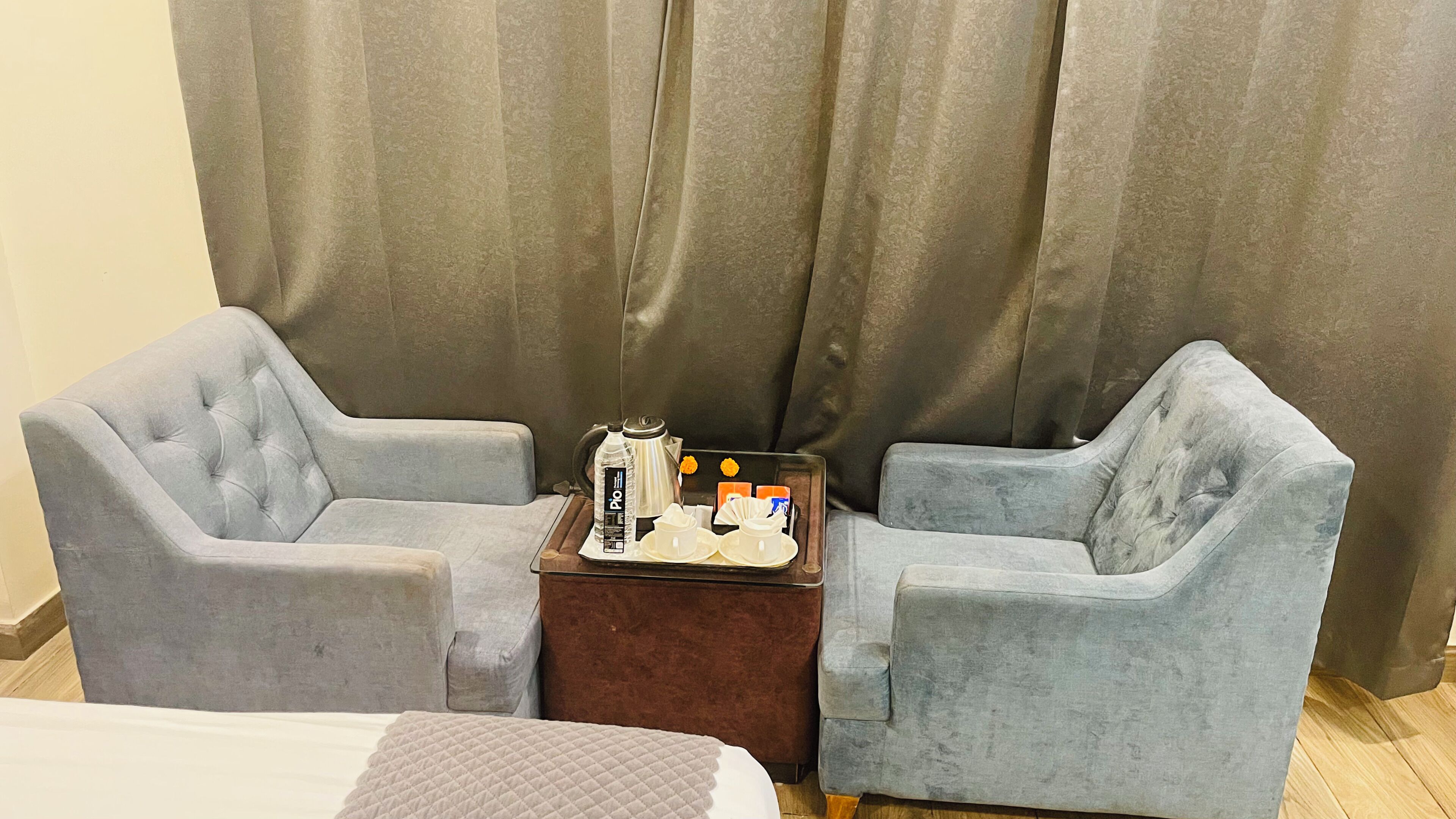 A room with two grey armchairs, a small wooden side table with a tray of toiletries, and a bed with a quilted cover.