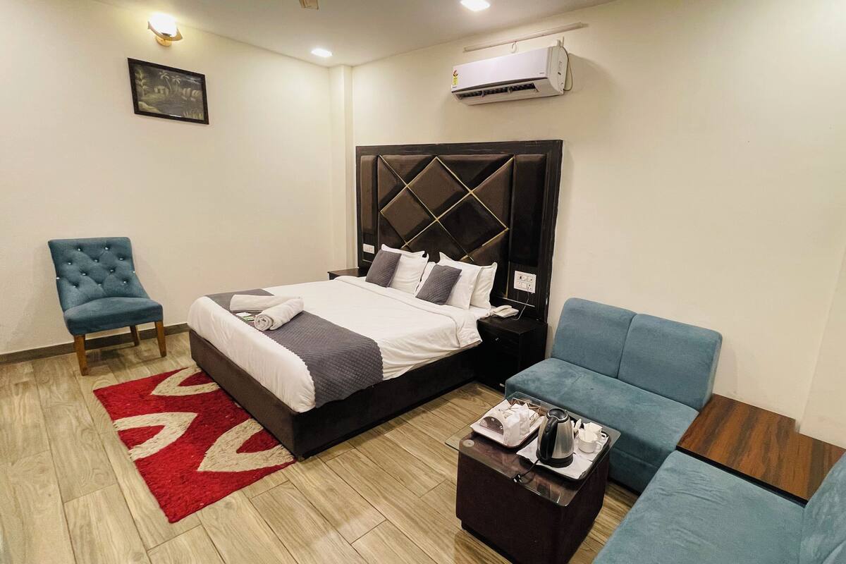 Una habitación de hotel con una cama, un sillón azul, un sofá, un tapete rojo y beige, una mesa de centro y un equipo de aire acondicionado.