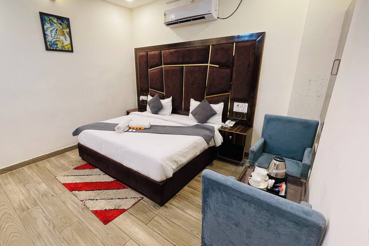 Una habitación de hotel con una cama, un sillón azul, una mesita con una taza y un plato, y un equipo de aire acondicionado en la pared.