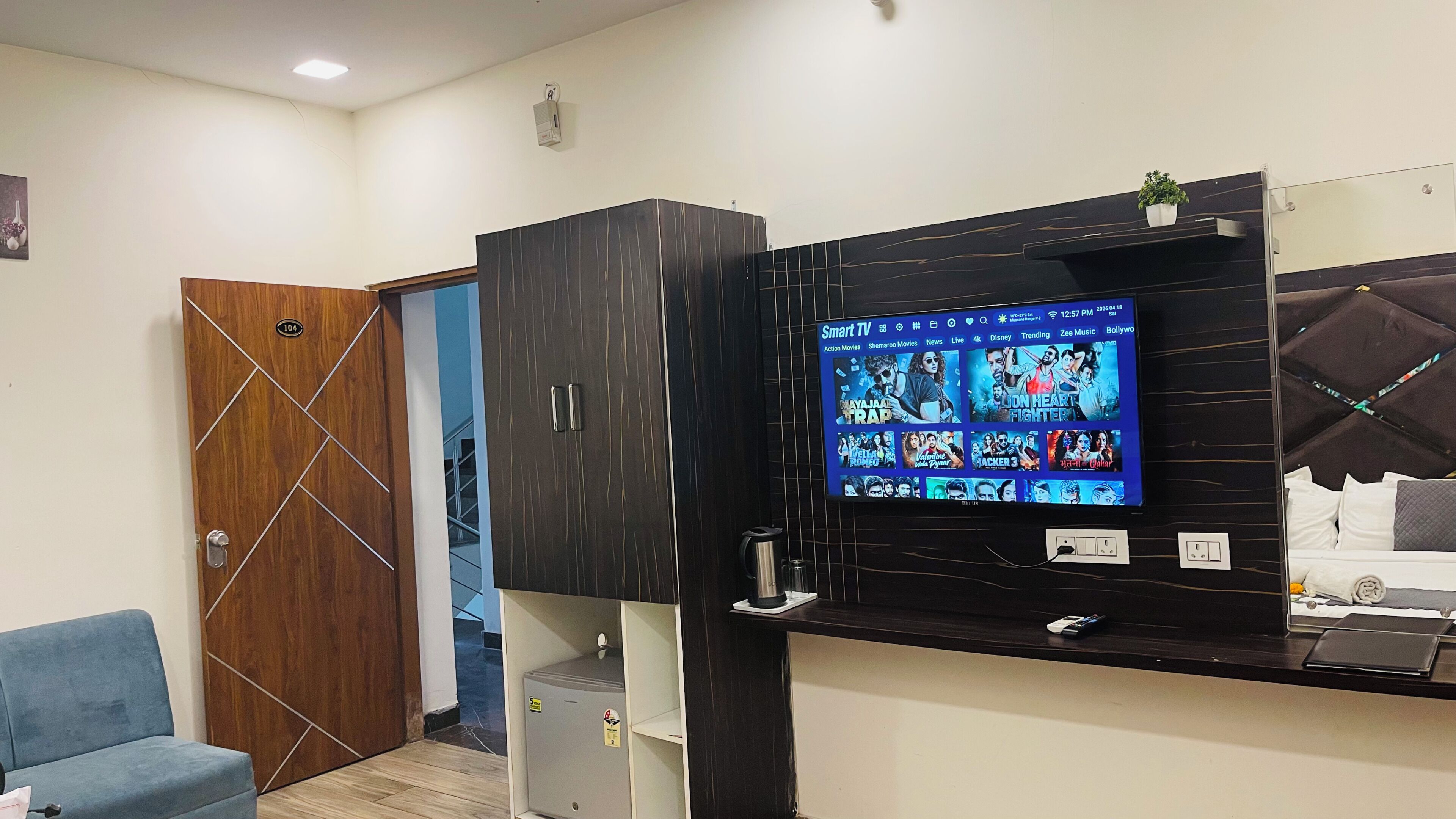 A hotel room with a TV displaying movie options, a wooden door, a blue chair, and a minibar.