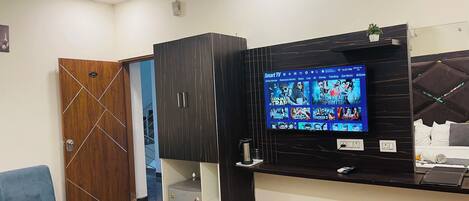 A hotel room with a TV displaying movie options, a wooden door, a blue chair, and a minibar.