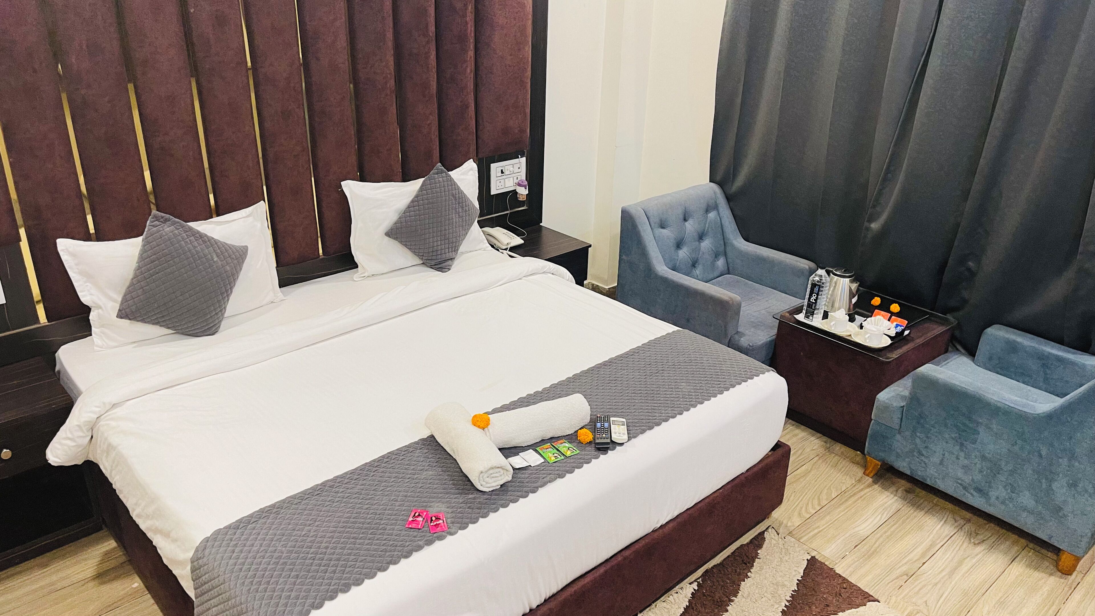 A hotel room with a large bed, a bedside table with a phone and a water bottle, a blue armchair, and a nightstand.