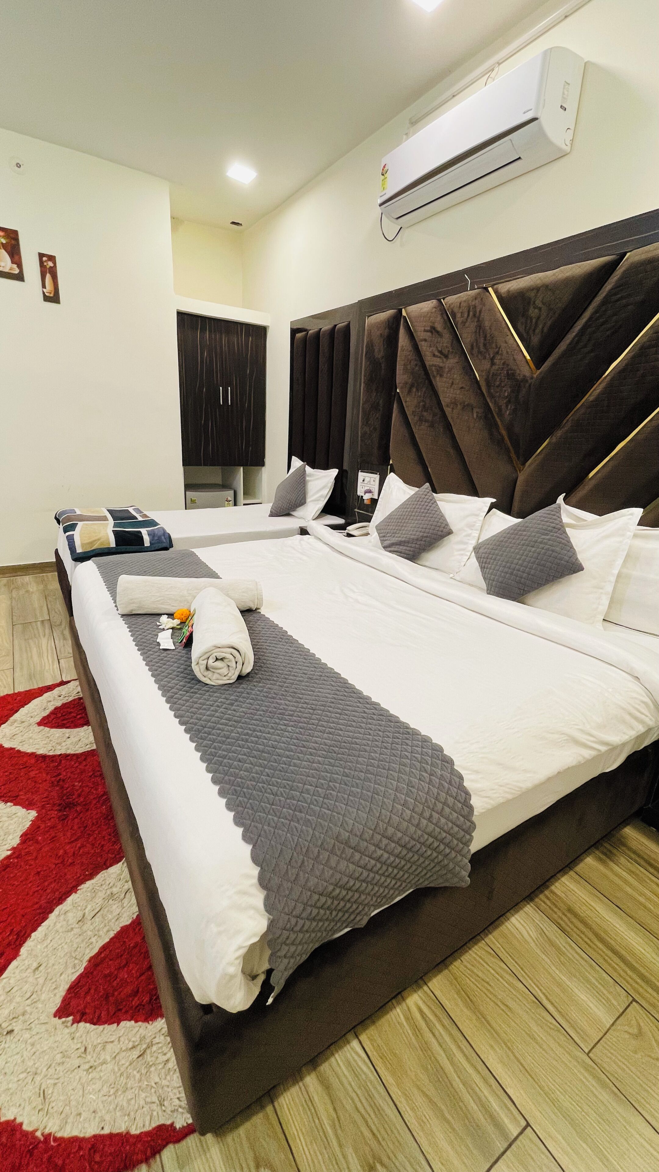 A modern hotel room with a large bed, a red and white rug, a wall-mounted air conditioner, and a dark headboard.