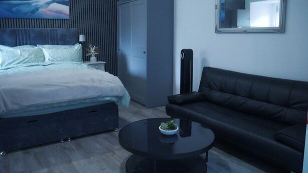 Ett modernt sovrum med en säng, en svart lädersoffa, ett runt soffbord med en växt, en garderob och en stor tavla på väggen.