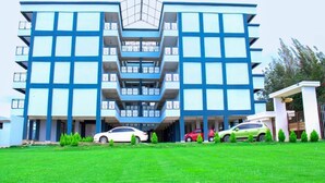 Un edificio moderno de varios pisos con un atrio central, múltiples balcones y un estacionamiento con varios automóviles.