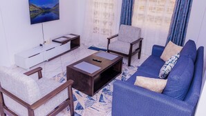 Una sala de estar con un televisor montado en la pared, un sofá, dos sillones, una mesa de centro y un mueble blanco para entretenimiento.